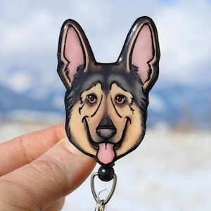 May include: A black and brown German Shepherd dog face is printed on a round, clear plastic badge holder. The badge holder is attached to a black and silver retractable badge reel.