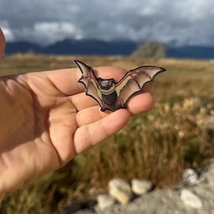 May include: A bat-shaped sticker with outstretched wings, featuring a tan body, black head, and reddish-brown wings. The sticker is held in a hand against a backdrop of a field and mountains. Text at the top reads "No AI used in my Art."