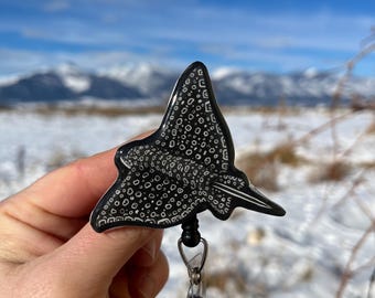 May include: A black and white spotted stingray design on a retractable badge holder. The holder has a clear plastic case and a black clip.