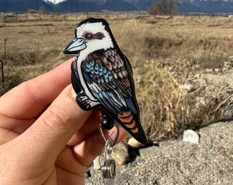 May include: A colorful badge reel featuring a detailed illustration of a kookaburra bird. The bird has a white head, blue beak, and brown, blue, and orange feathers. The text "No AI used in my Art" is visible. The reel has a black bead and a clear plastic clip.