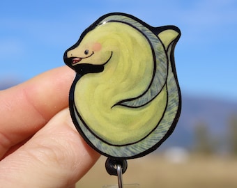 May include: A yellow and black badge reel featuring a cartoon eel design. The eel has a smiling face with pink cheeks and a red mouth. The badge reel has a black retractable cord and a clear plastic holder.