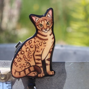 May include: A wooden cutout of a brown and black spotted cat sitting with its tail curled around its legs. The cat has green eyes and a light brown nose.
