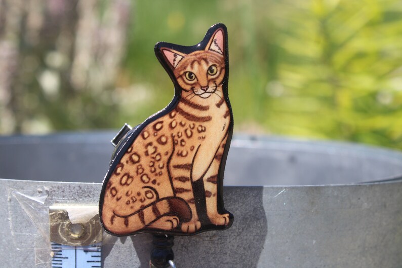 May include: A wooden cutout of a brown and black spotted cat sitting with its tail curled around its legs. The cat has green eyes and a light brown nose.