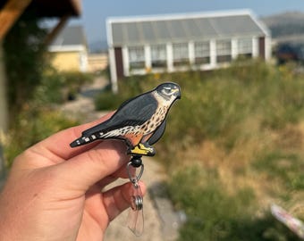 May include: A badge reel featuring a detailed illustration of a bird of prey, with black, white, and orange markings. The bird is attached to a clear plastic reel with a metal clip.