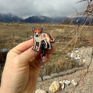 May include: A red and white cartoon dog badge reel with a black clip. The dog is standing on all four legs and has a black nose and eyes. The background is a blurry image of a field and mountains.