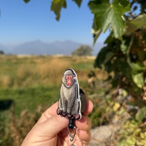 May include: A badge reel featuring a detailed illustration of a Japanese macaque monkey. The monkey is depicted in shades of gray with a red face, sitting upright. The reel has a black retractable cord and a clear plastic ID holder.