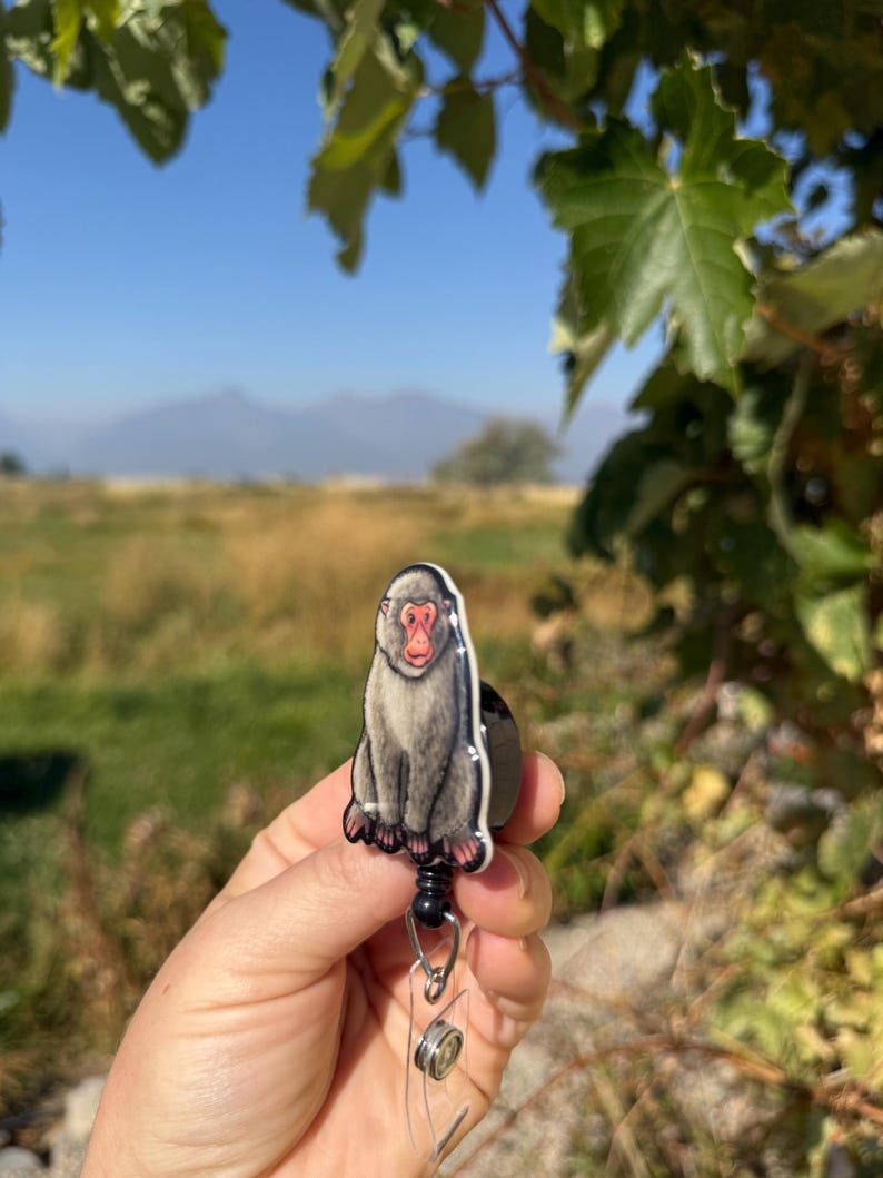 May include: A badge reel featuring a detailed illustration of a Japanese macaque monkey. The monkey is depicted in shades of gray with a red face, sitting upright. The reel has a black retractable cord and a clear plastic ID holder.