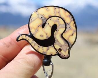 May include: A yellow and black patterned snake curled up in a circle, with a black and white eye, on a retractable badge holder.