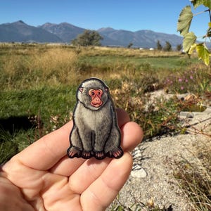 May include: A gray and black monkey sticker with a red face is held in a hand. The sticker is in front of a scenic background of mountains, fields, and a blue sky. The monkey is sitting upright.