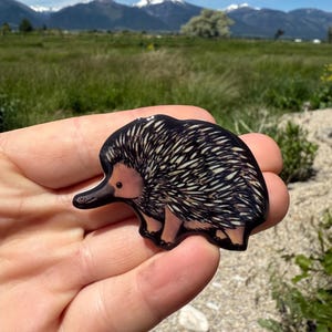 May include: A small, flat, decorative pin shaped like an echidna, featuring a black and tan color palette with detailed quill-like markings on its back, a long snout, and a smooth, glossy finish. The text 'No AI used in my Art' is visible.