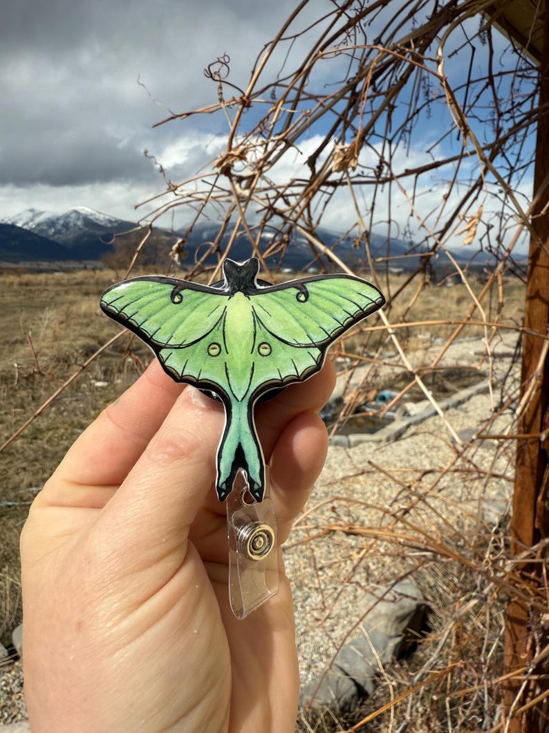 Puede incluir: Un carrete de insignia de polilla luna verde y negro con un clip de pl&aacute;stico transparente. La polilla tiene una cola larga y se representa con detalles realistas.