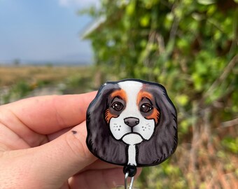 May include: A black and white cartoon dog with brown markings on its face is printed on a round badge holder. The badge holder is attached to a retractable badge reel with a clear plastic strap.