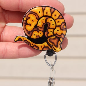 May include: A yellow and brown snake curled up on a black background. The snake is on a retractable badge holder with a black bead and a silver clip.