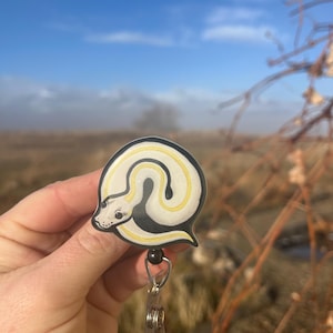 May include: A white and yellow snake with black markings is curled up on a black and white badge holder. The badge holder has a clear plastic clip and a metal retractable reel.