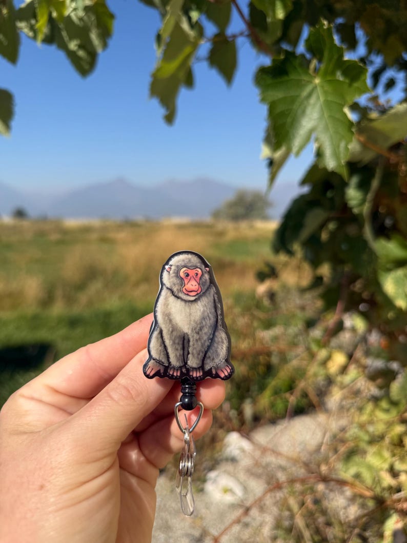 May include: A badge reel featuring a cartoon illustration of a Japanese macaque monkey. The monkey has gray fur, a red face, and is sitting upright. The reel has a black retractable cord and a metal clip.