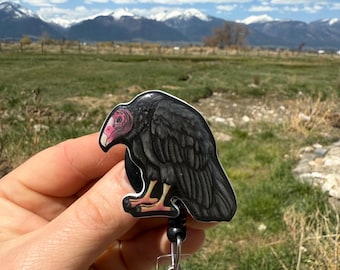 May include: A black and white cartoon image of a black vulture with a pink face on a black retractable badge holder with a silver clip.