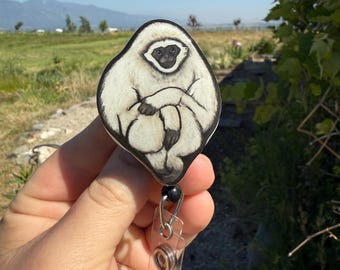 May include: A white and black badge reel featuring a cartoon gibbon illustration. The gibbon is depicted with its arms crossed. The badge reel has a clear plastic clip and a black bead. The background is a natural outdoor scene.