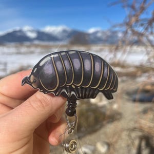 May include: A black and purple pill bug with a detailed illustration, attached to a black and silver retractable badge reel.