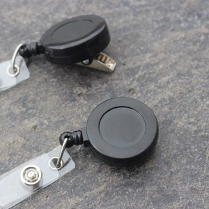 May include: Two black retractable badge holders with clear plastic badge clips. The badge holders have a metal clip on one side and a retractable cord on the other.