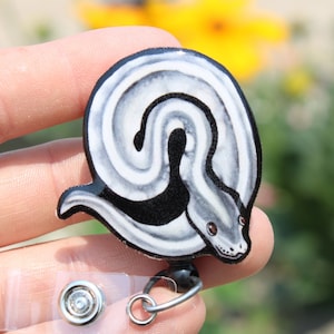 May include: A round, black and white snake design badge reel. The snake is depicted in a swirling pattern with a black underbelly and a detailed head with brown eyes. The reel has a clear plastic strap and a metal clip.