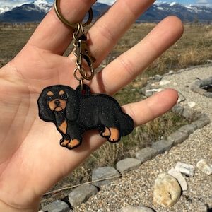 May include: Black and tan Cavalier King Charles Spaniel keychain. The dog-shaped charm is attached to a gold-colored keyring and clasp. The keychain is held up against a backdrop of mountains and a blue sky.