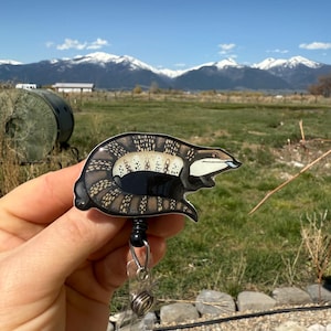 May include: A black and white illustrated blue-tongued skink on a retractable badge reel. The skink is curled up with its tongue sticking out. The background is a blurry image of a mountain range.