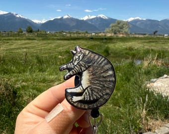 May include: A badge reel featuring a sleeping tabby cat design. The cat is depicted in shades of brown and gray with black stripes. The reel has a black cord and a clear plastic clip.