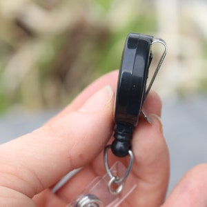 May include: Black retractable badge holder with a metal clip and a clear plastic badge holder. The badge holder is attached to a silver chain.