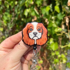 May include: A retractable badge holder with a round, black plastic casing. The front of the casing features a hand-painted image of a Cavalier King Charles Spaniel dog with brown, white, and orange fur.