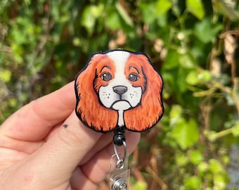 May include: A retractable badge holder with a round, black plastic casing. The front of the casing features a hand-painted image of a Cavalier King Charles Spaniel dog with brown, white, and orange fur.