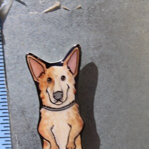 May include: A hand-painted ceramic tile featuring a cartoon dog with brown, white, and black markings. The dog is wearing a black collar and has a friendly expression.