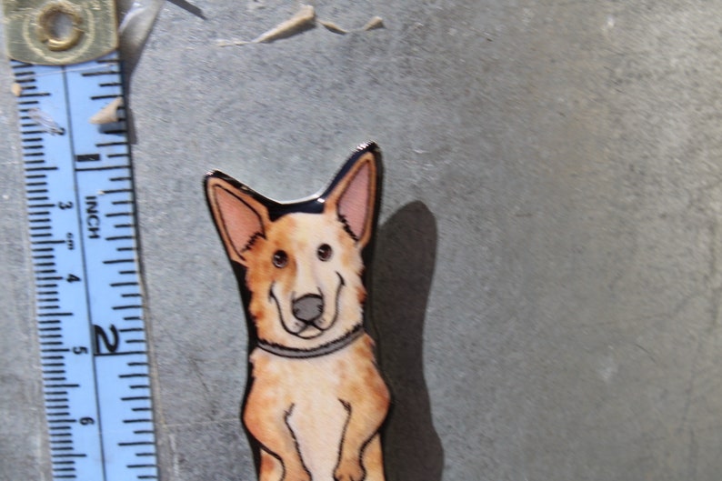 May include: A hand-painted ceramic tile featuring a cartoon dog with brown, white, and black markings. The dog is wearing a black collar and has a friendly expression.