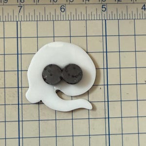 May include: A white, abstract-shaped magnet with two dark gray circular magnets. The magnet has a curved, flowing design with a tail-like extension. The object is placed on a grid-lined surface.