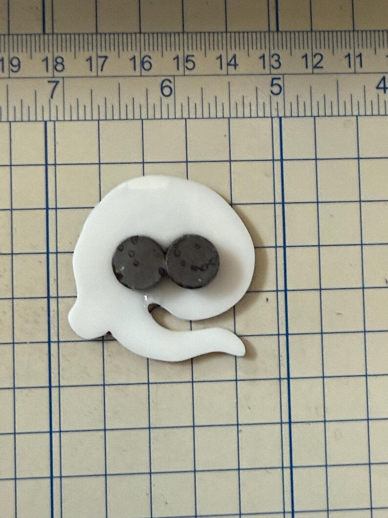 May include: A white, abstract-shaped magnet with two dark gray circular magnets. The magnet has a curved, flowing design with a tail-like extension. The object is placed on a grid-lined surface.