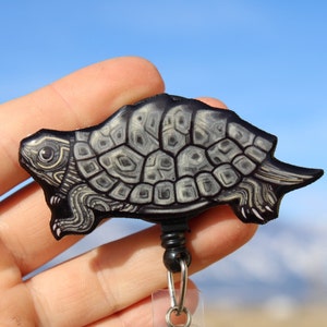 May include: A black and white turtle-shaped retractable badge holder with a detailed design. The turtle is holding a black retractable cord.