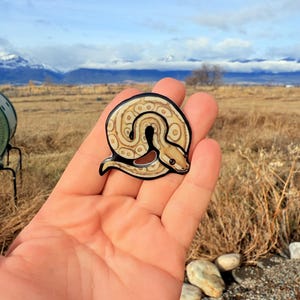 May include: A circular snake-shaped sticker with a black border. The snake has a tan and brown pattern with a brown patch. The sticker is held in a hand against a backdrop of a field, mountains, and a cloudy sky.