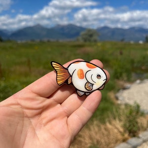May include: A white and orange hand-painted fish pin with black outlines. The fish has a round body and large, round eyes. The pin is a cute and whimsical accessory.