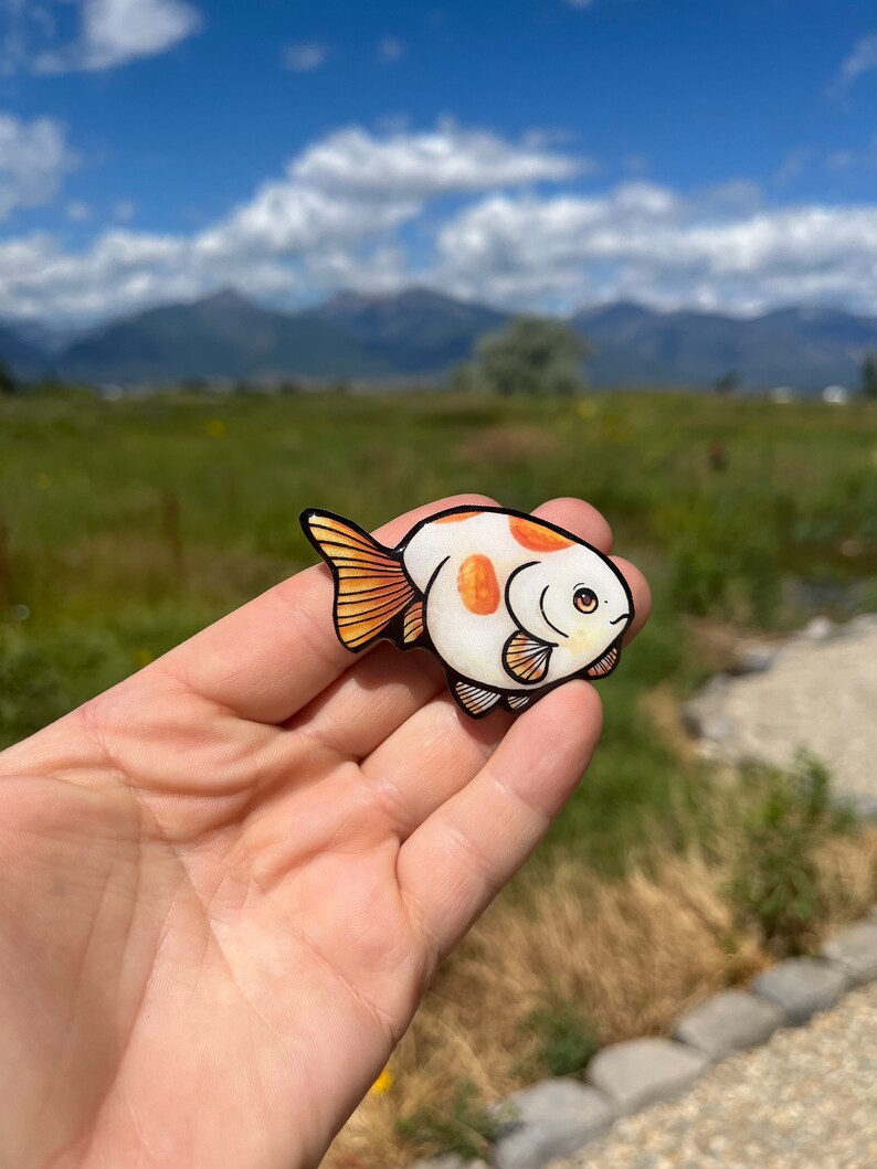 May include: A white and orange hand-painted fish pin with black outlines. The fish has a round body and large, round eyes. The pin is a cute and whimsical accessory.