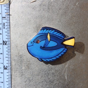 May include: A blue and yellow painted wooden fish figurine. The fish has a black eye and a yellow fin. The fish is about 2 inches long.