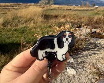 May include: A black, white, and tan Cavalier King Charles Spaniel badge reel. The badge reel has a retractable cord and a clear plastic holder. The text "No AI used in my Art" is in the upper left corner.