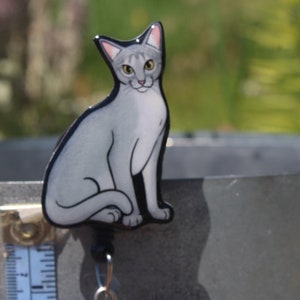May include: A gray and white cat figurine with a black outline. The cat is sitting with its tail curled around its legs. The figurine is attached to a retractable badge reel with a silver metal clip.