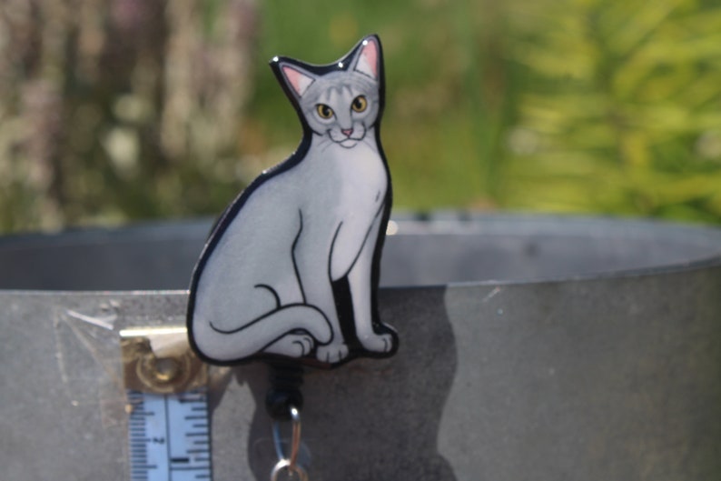 May include: A gray and white cat figurine with a black outline. The cat is sitting with its tail curled around its legs. The figurine is attached to a retractable badge reel with a silver metal clip.