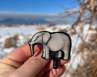 May include: A gray elephant-shaped retractable badge holder with a black swivel clip and a clear plastic badge holder. The badge holder is in a hand against a snowy background.