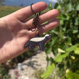 May include: A wooden keychain featuring a badger design. The badger is black and white with a small, reddish-brown nose. The keychain has a bronze-colored clasp and ring. The background shows a blurred view of green foliage and a mountain range.