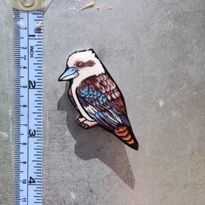 Op de afbeelding: Een houten uitsnijding van een kookaburra met blauwe, bruine en witte veren. De vogel is ongeveer 5 cm hoog en zit op een metalen oppervlak.
