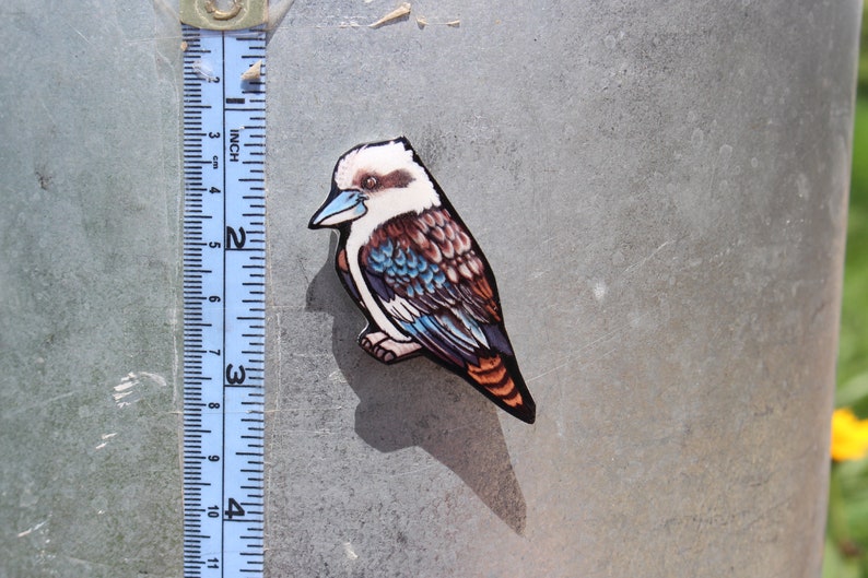 Op de afbeelding: Een houten uitsnijding van een kookaburra met blauwe, bruine en witte veren. De vogel is ongeveer 5 cm hoog en zit op een metalen oppervlak.