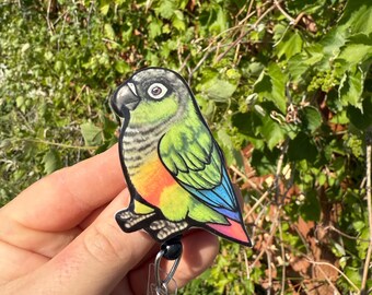 May include: A colorful parrot badge reel featuring a green, yellow, blue, and red bird design. The badge reel has a black retractable cord and a clear plastic clip for attaching to clothing or accessories. The background is a blurred view of green foliage.