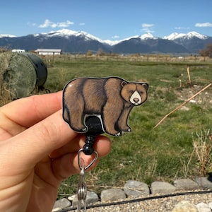 May include: A brown bear badge reel with a black retractable cord and a silver swivel clip. The bear is facing the left side of the image. The background is a blurry image of a mountain range.