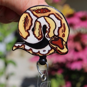 May include: A yellow, white, and brown snake with black markings is printed on a plastic badge holder. The badge holder has a black swivel clip and a clear plastic strap.
