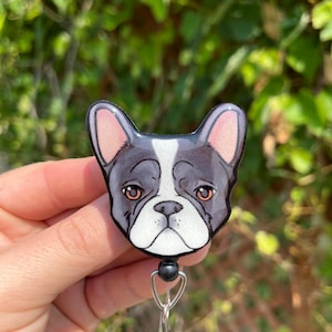 May include: A black and white cartoon French bulldog face on a retractable badge holder with a black clip and a silver chain.